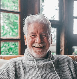 An older, smiling man wearing a sweater and sitting on a sofa